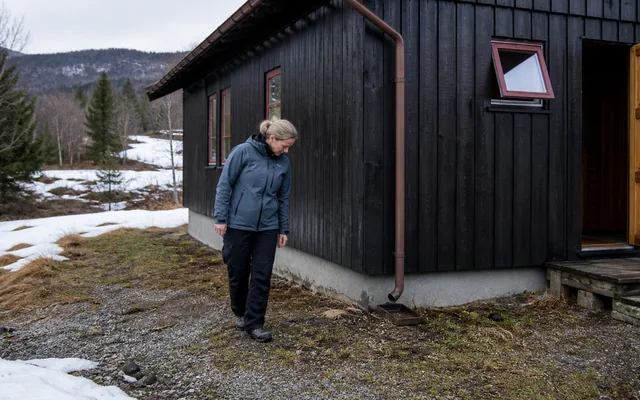 Hytteeier undersøker bakken og taknedløpet ved en mørk trehytte under vårens snøsmelting.